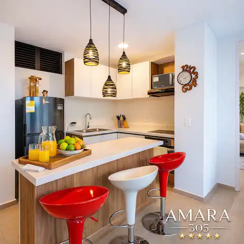 Cocina americana con barra, tres lámparas colgantes, tres taburetes (dos rojos, uno blanco) y bandeja con frutas y jugo.